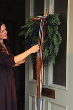 A woman in a dark plum dress is adjusting a large holiday wreath hanging on a white paneled door. The wreath is made of green evergreen branches, decorated with dark blue berries, and tied with a long brown ribbon with blue and white stripes.