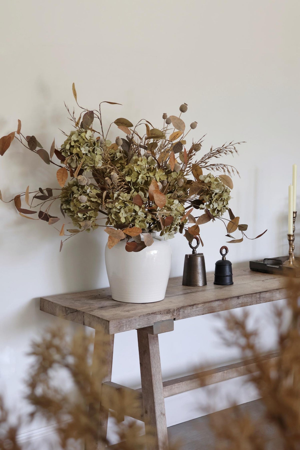 White vase with faux dried flowers on a wooden table against a white wall
