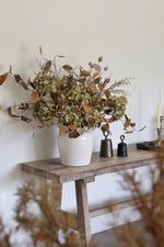 White vase with faux dried flowers on a wooden table against a white wall