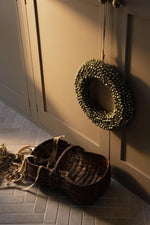 brass bell wreath hanging on soft brown cupabord doors with a vintage woven basket full of ribbons on the tiled floor 
