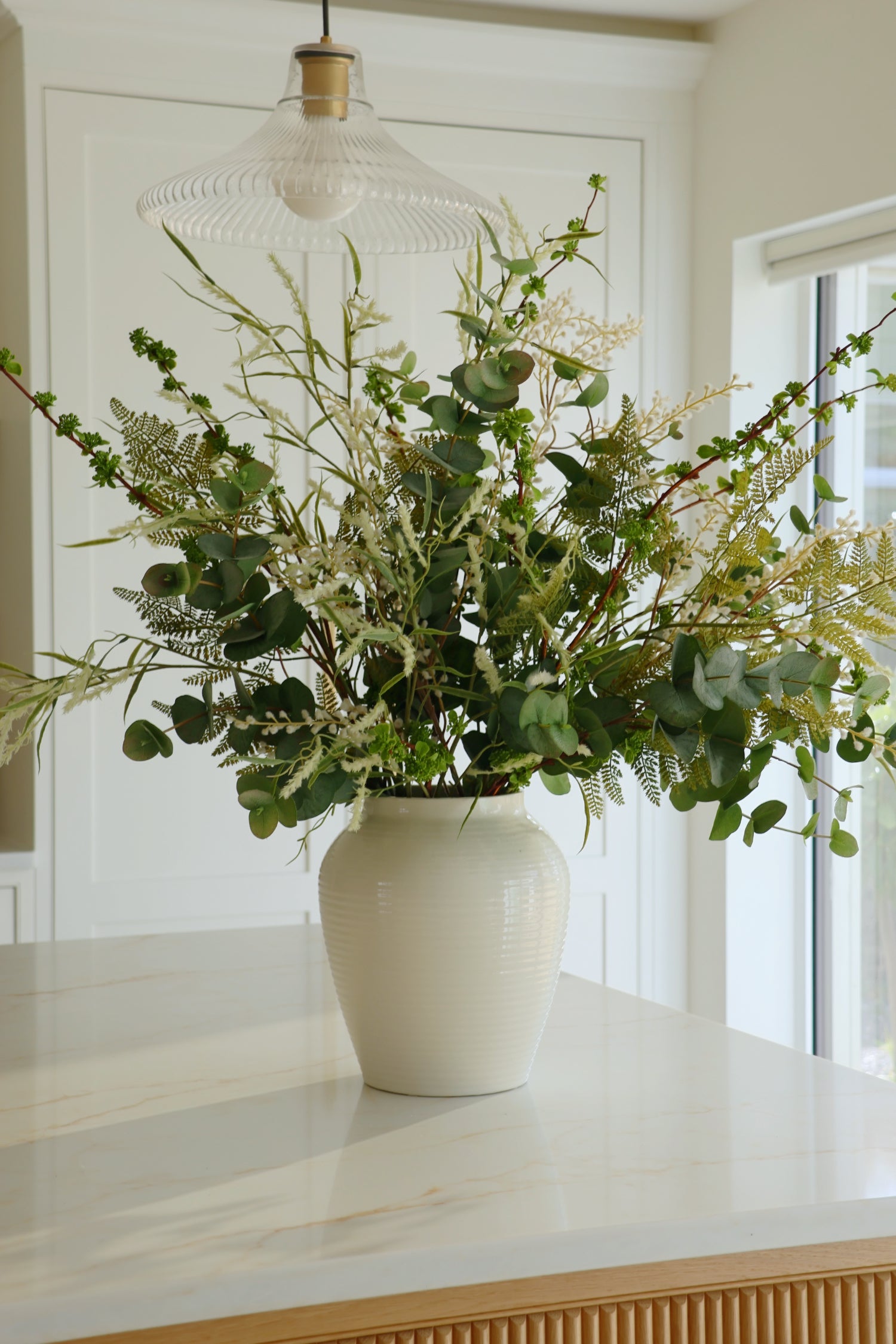 White vase with greenery on a white surface in a bright room