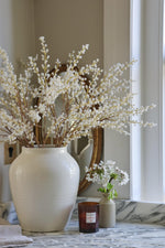 A collection of artificial white willow stems arranged in a white ceramic vase on a marble surface, with a candle and a small pot with real flowers in the background.