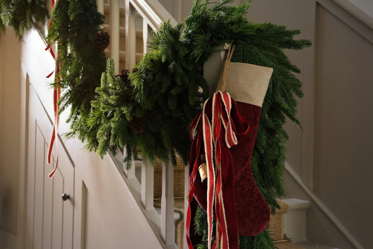 A green Christmas wreath with pinecones and ribbons hangs on a white staircase railing next to a red velvet stocking with cream trim and long red and cream ribbons.