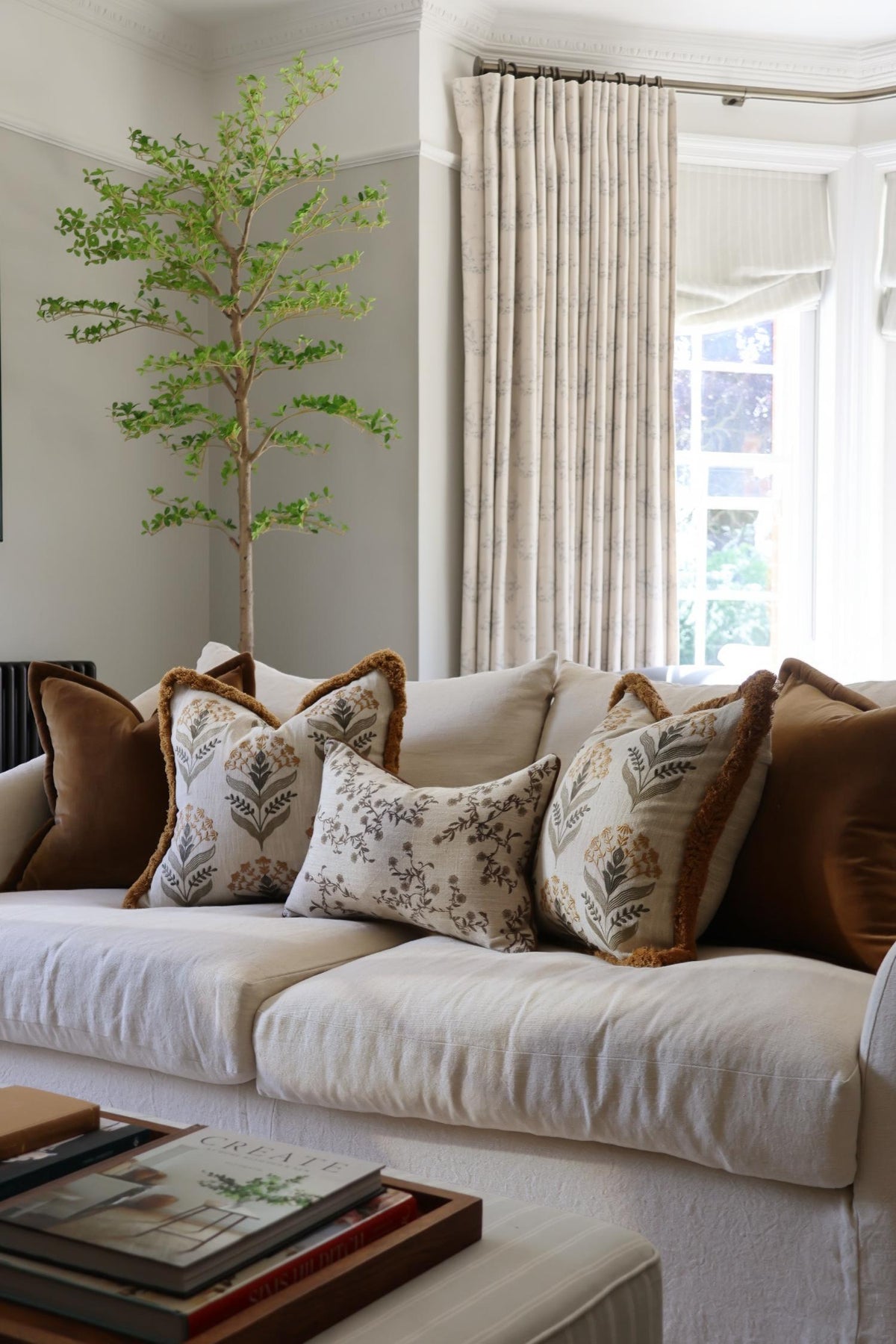 Decorative cushions on a beige sofa in a living room with a tree and window in the background.