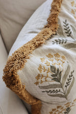 Decorative cushion with large yellow fringe trim and embroidered floral patterns on a neutral background