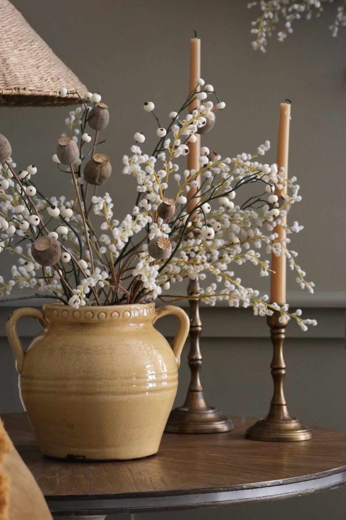 a yellow ceramic pot with handles and artificial white foliage in on a wooden side table with brass candlesticks behind it