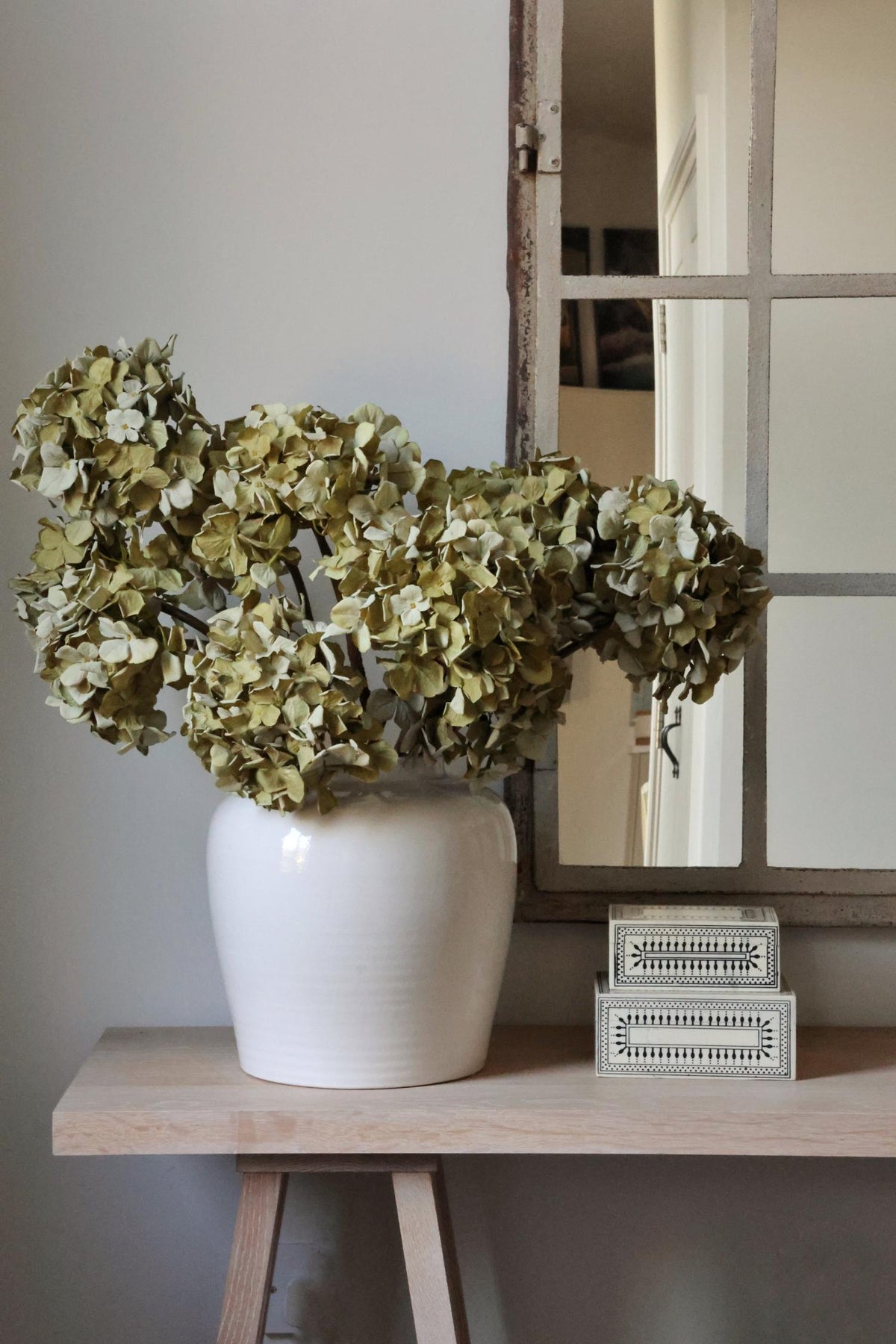 White vase with faux dried moss green hydrangeas on a wooden table next to a mirror