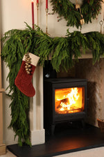 logburner dressed in draped faux norfolk pine garlands and a red stocking with brass bells in cosy lounge setting