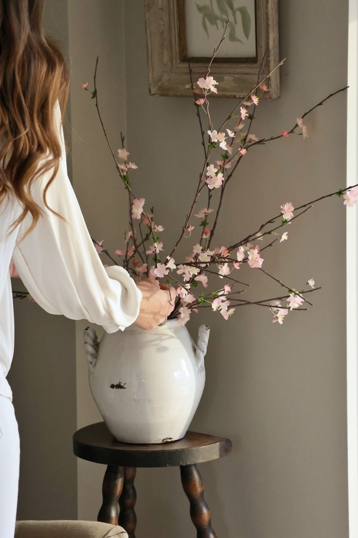 White vase on wooden stool with pink blossom branches being arranged by ladies arm