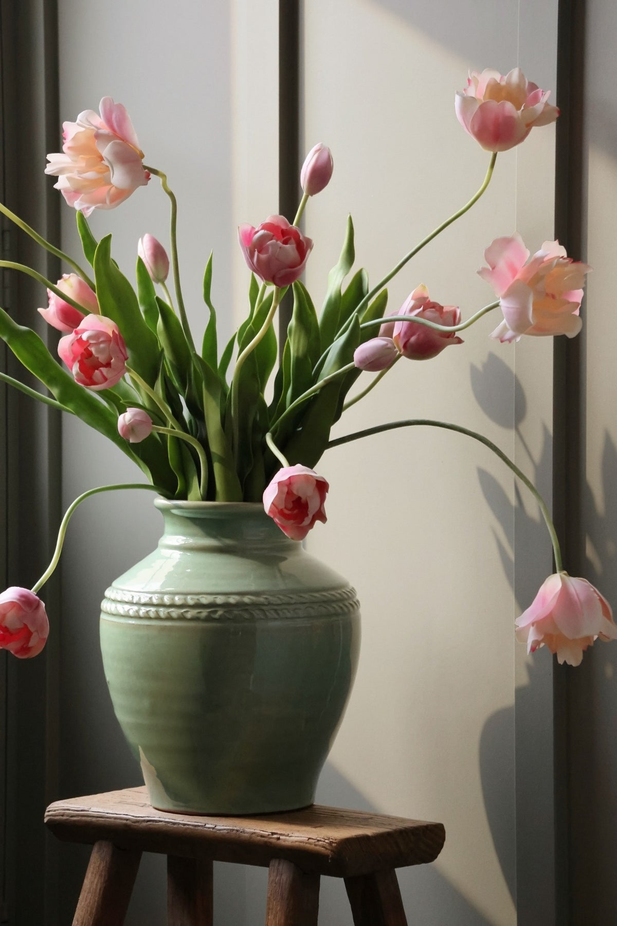 Green vase with pink tulips on a wooden stool against a neutral wall