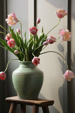 Green vase with pink tulips on a wooden stool against a neutral wall