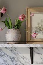 Pink tulips in a white vase on a marble surface with a framed picture in the background.