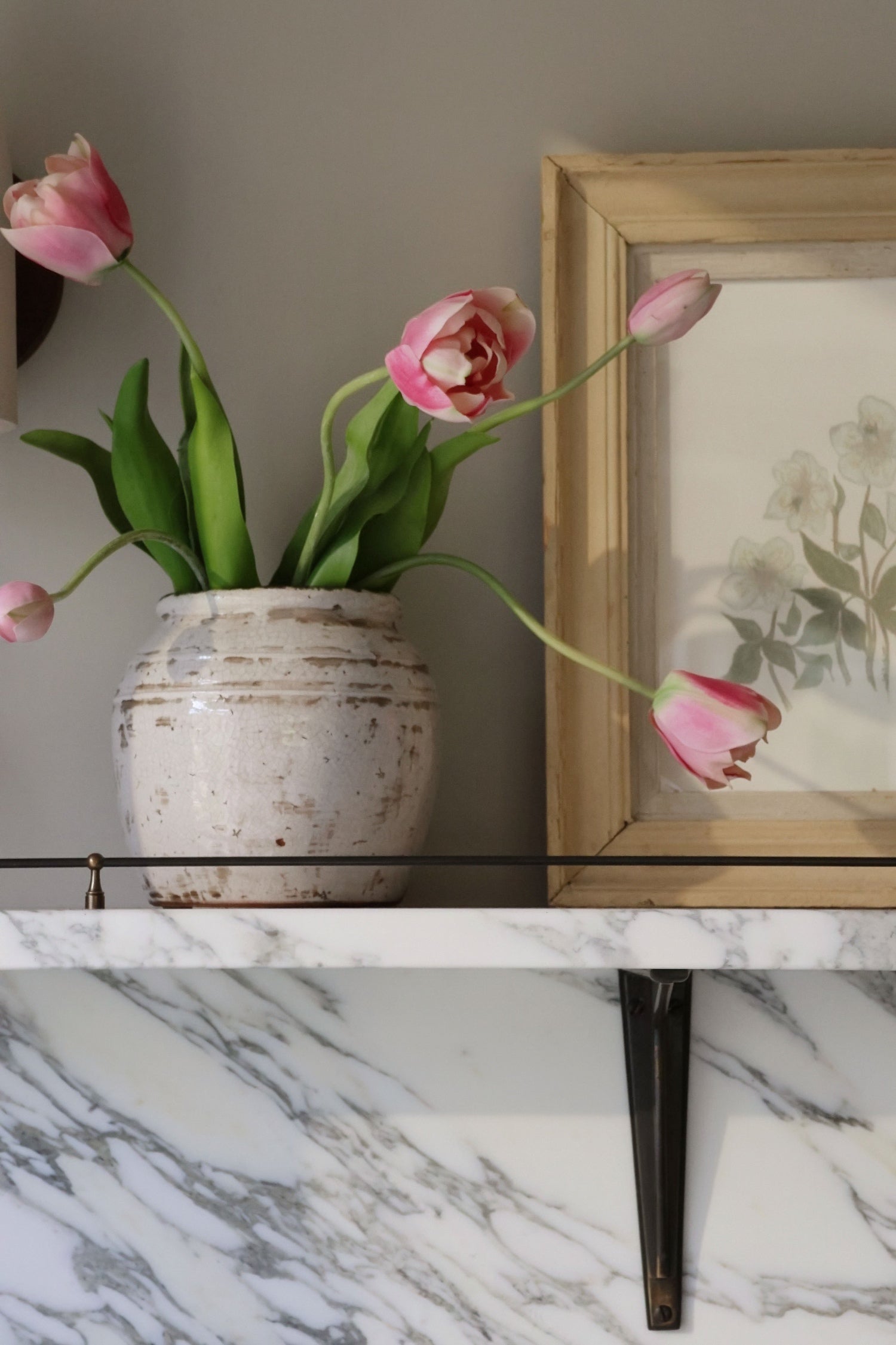Pink tulips in a white vase on a marble surface with a framed picture in the background.