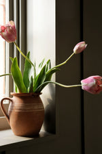 Pink tulips in a terracotta pot on a windowsill with a dark wooden cabinet in the background.