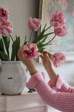 pink tulips in white vase being arrangement by womens hands in pink jumper on mantle