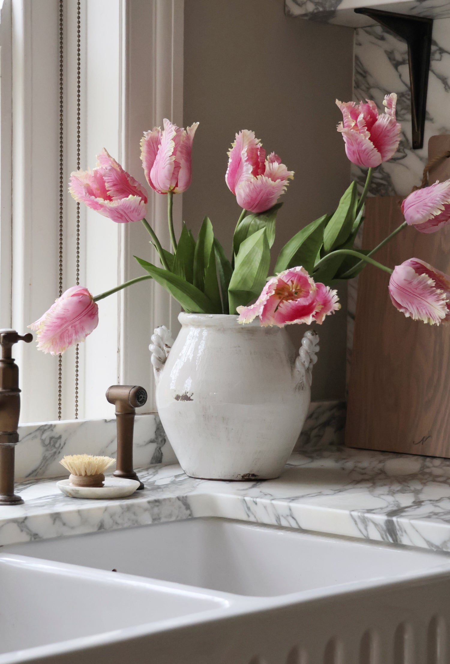Pink tulips in a white vase on a marble countertop with a window in the background.