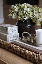 A styled interior scene showing a dark brown ceramic vase filled with pale green faux hydrangea blooms, placed on a woven tray beside stacked white decorative boxes, a silver tray with a small brass bell, and a white candle.