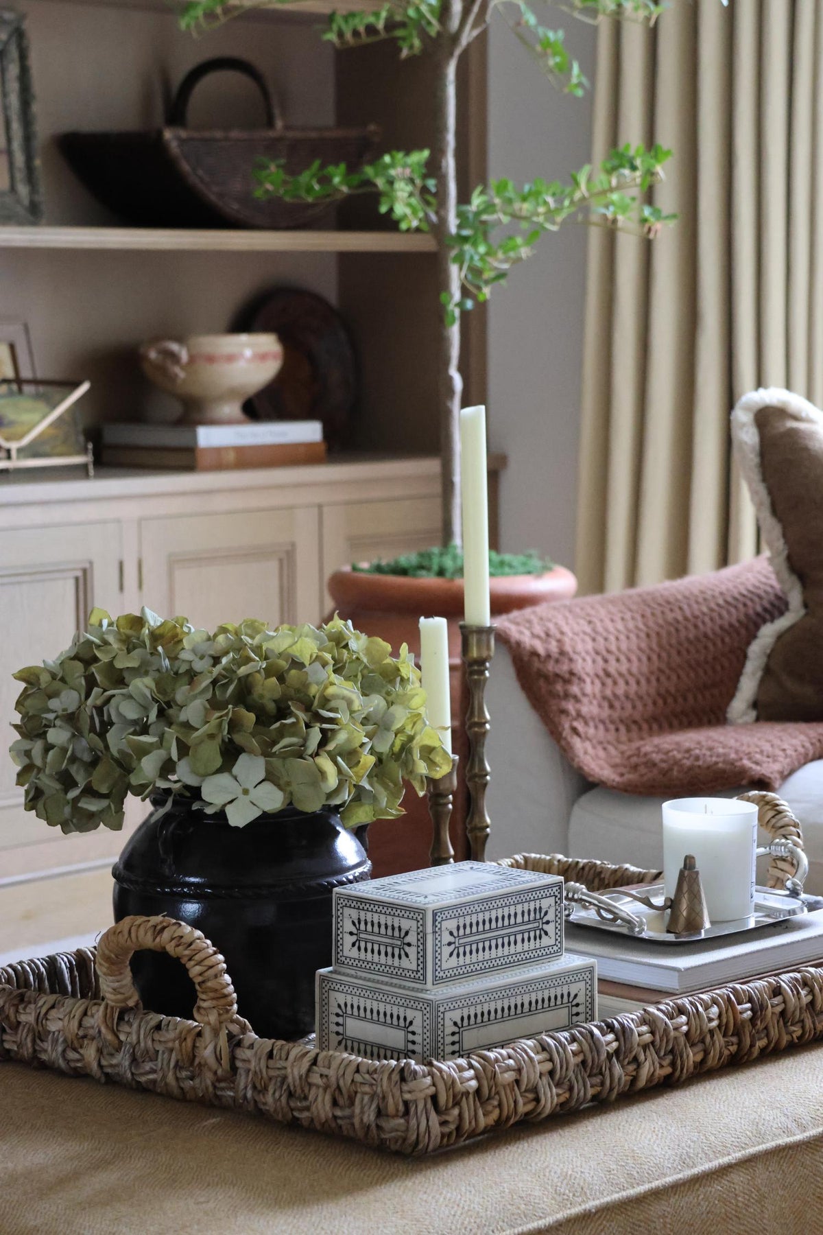 A styled home decor scene showing a woven rectangular tray holding a black vase with green faux hydrangeas, two stacked resin decorative boxes, white candles, and a small plant, set in a living room with a sofa and shelving in the background.
