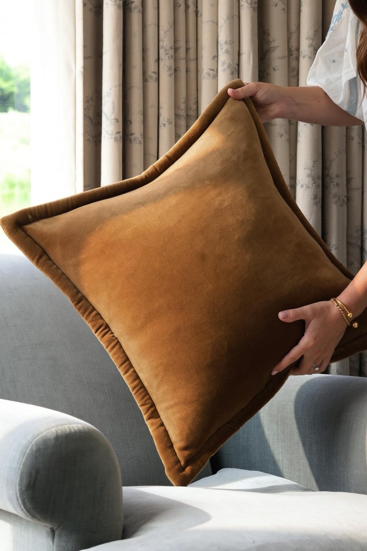 Brown rcushion being held above a gray sofa with curtains in the background