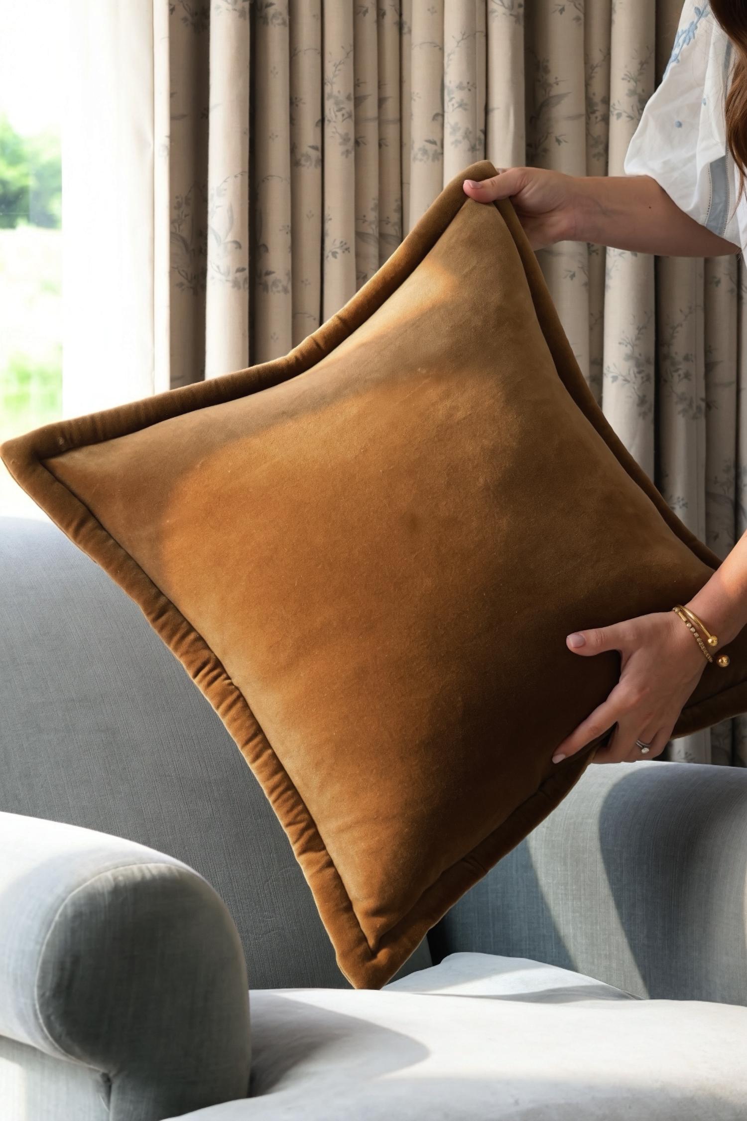 Brown rcushion being held above a gray sofa with curtains in the background