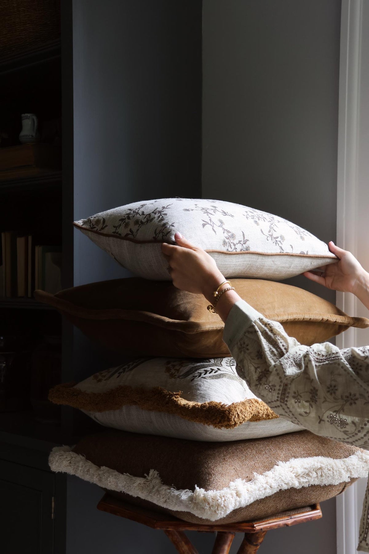 Person arranging decorative cushions on a wooden stool against a neutral wall.