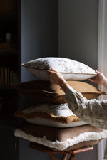 Person arranging decorative cushions on a wooden stool against a neutral wall.