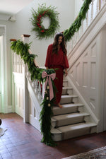 Woman in red dress standing on a staircase decorated with artificial christmas garlands and a large wreath with red berries.