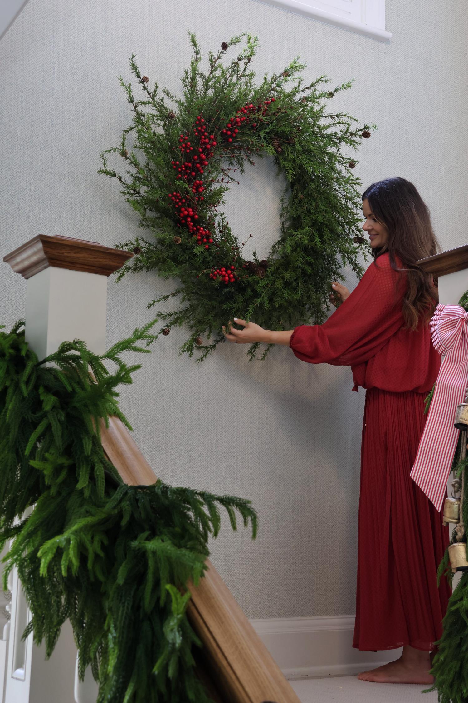 Woman in a red dress hanging a faux large wreath with red berries on a staircase wall.