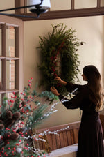 Woman arranging a faux wreath on a door with festive decorations.