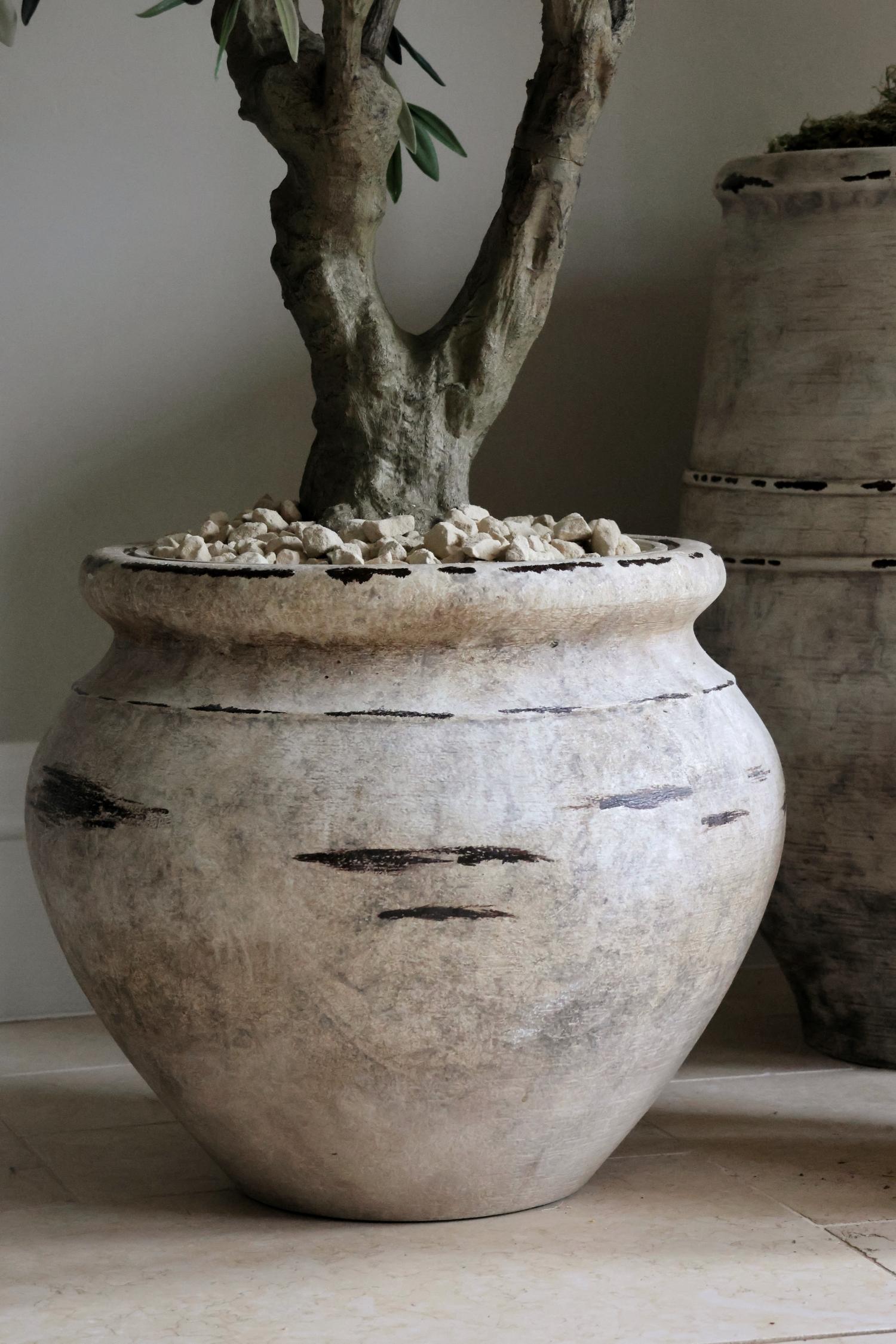 A lightweight, faux barrow planter made to look like weathered stone, containing a small tree. The planter is placed on a floor and has a rustic yet elegant appearance.