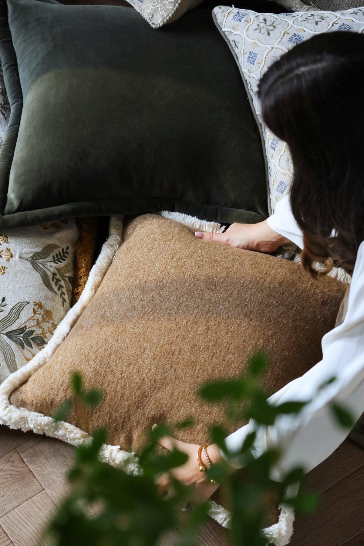 Person arranging cushions on wooden floor with plant in foreground