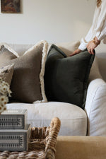 Person arranging dark green cushions on a sofa in a cozy living room.