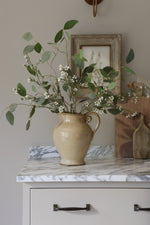 Beige ceramic pitcher with faux waxflower on a marble surface, framed picture in the background