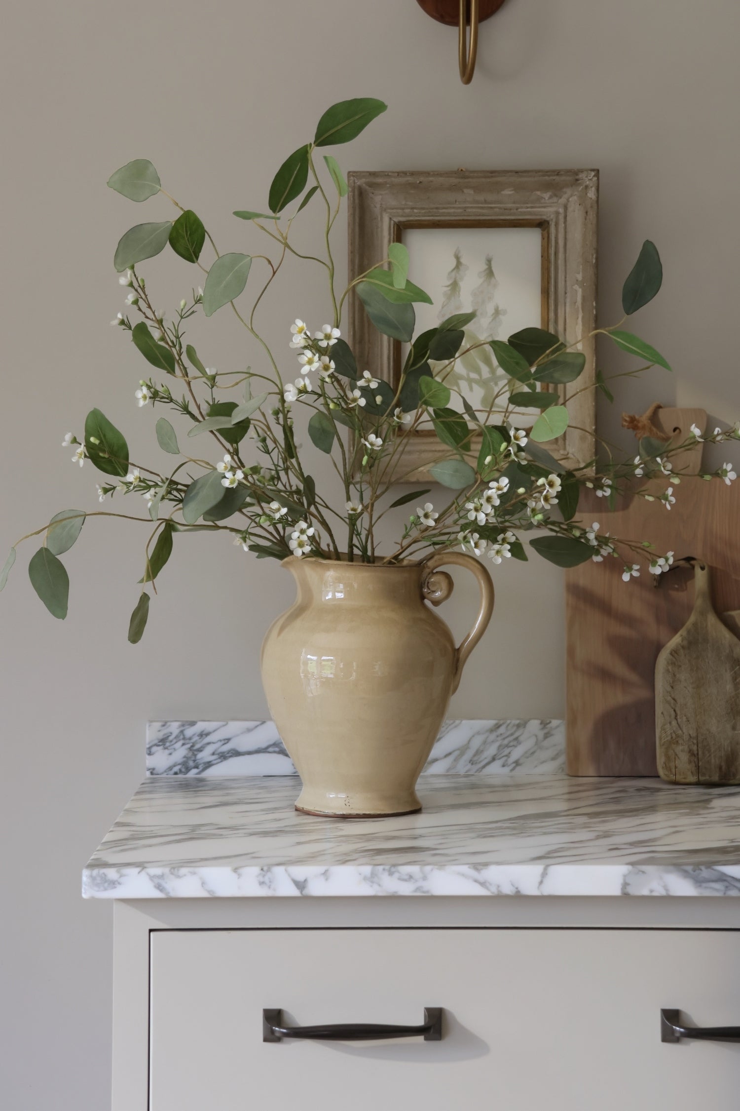 Beige ceramic pitcher with faux waxflower on a marble surface, framed picture in the background