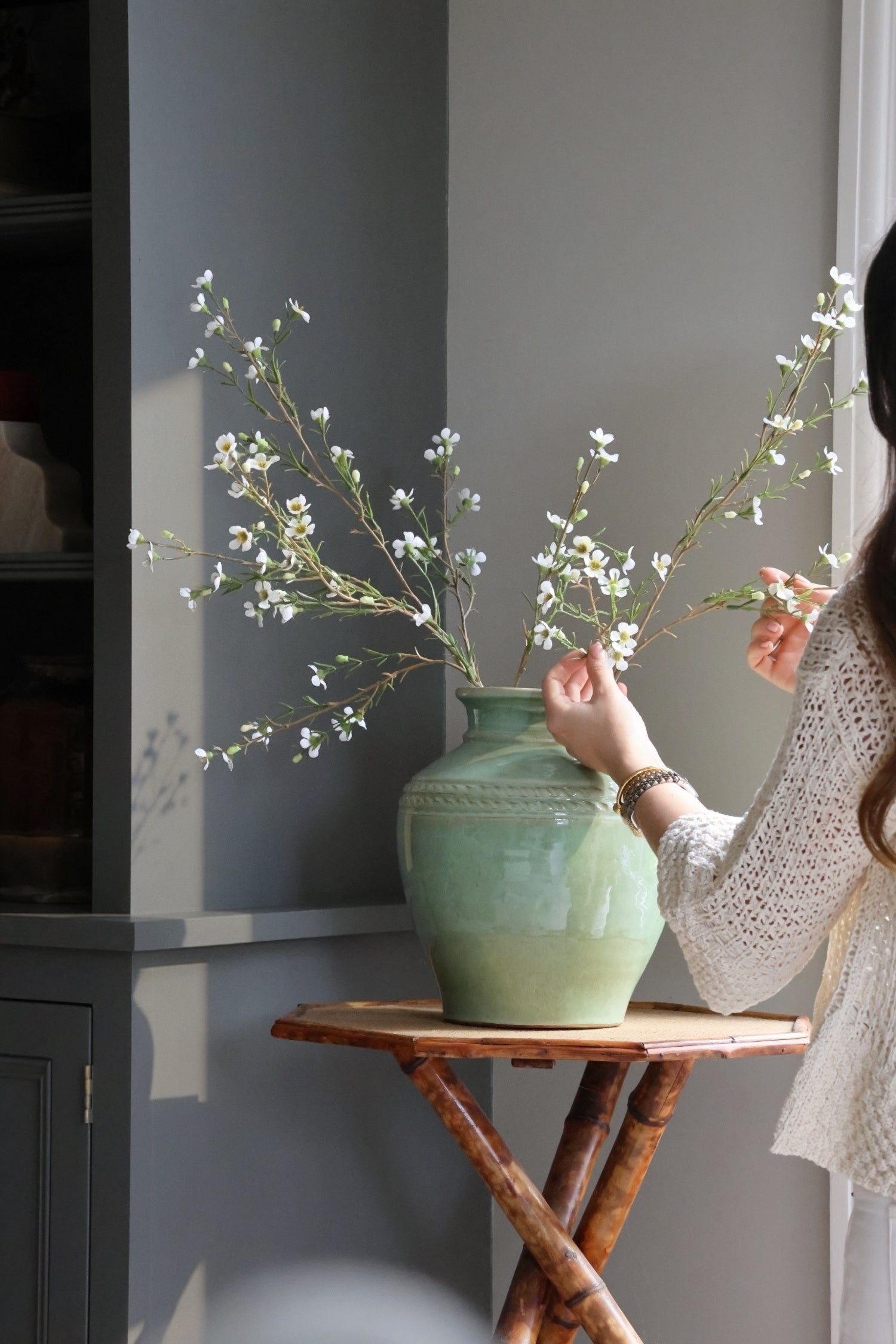 Person arranging white waxflower stems into a green vase on a small wooden table.