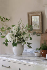 White vase with green and white flowers on a marble countertop with a framed picture in the background.
