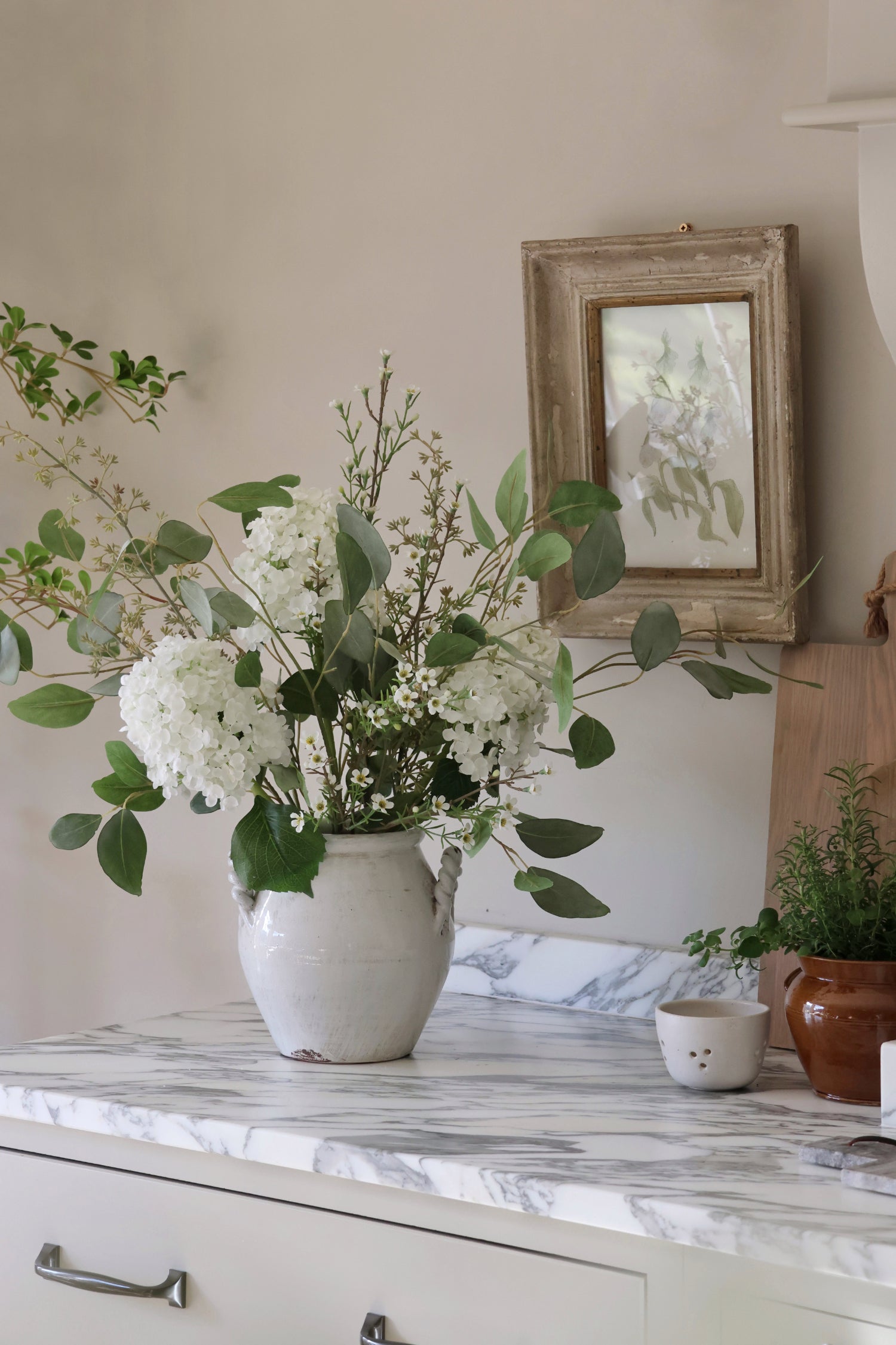 White vase with green and white flowers on a marble countertop with a framed picture in the background.