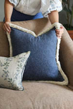 Person holding a blue cushion with beige fringe trim on a couch