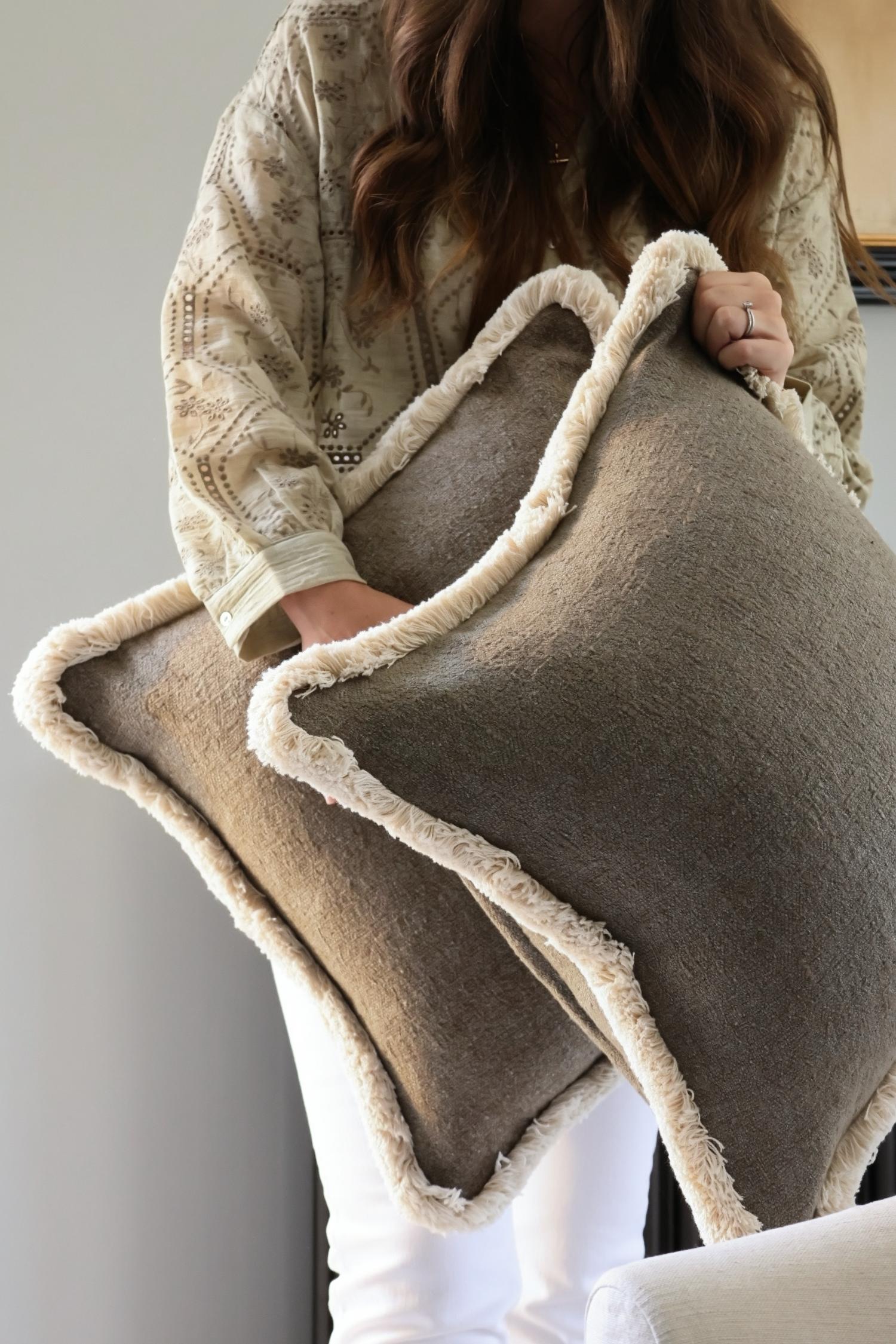 Person holding large green textured cushions with beige fringe trim against a neutral background