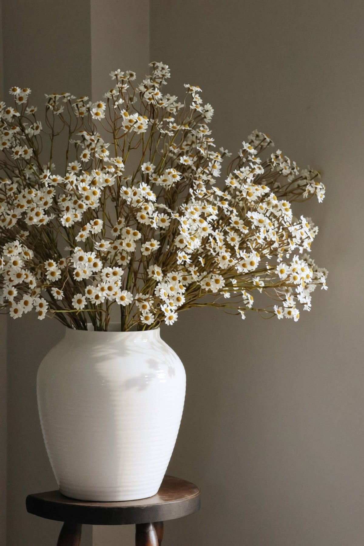 White vase with white daisyflowers on a wooden stool against a plain wall