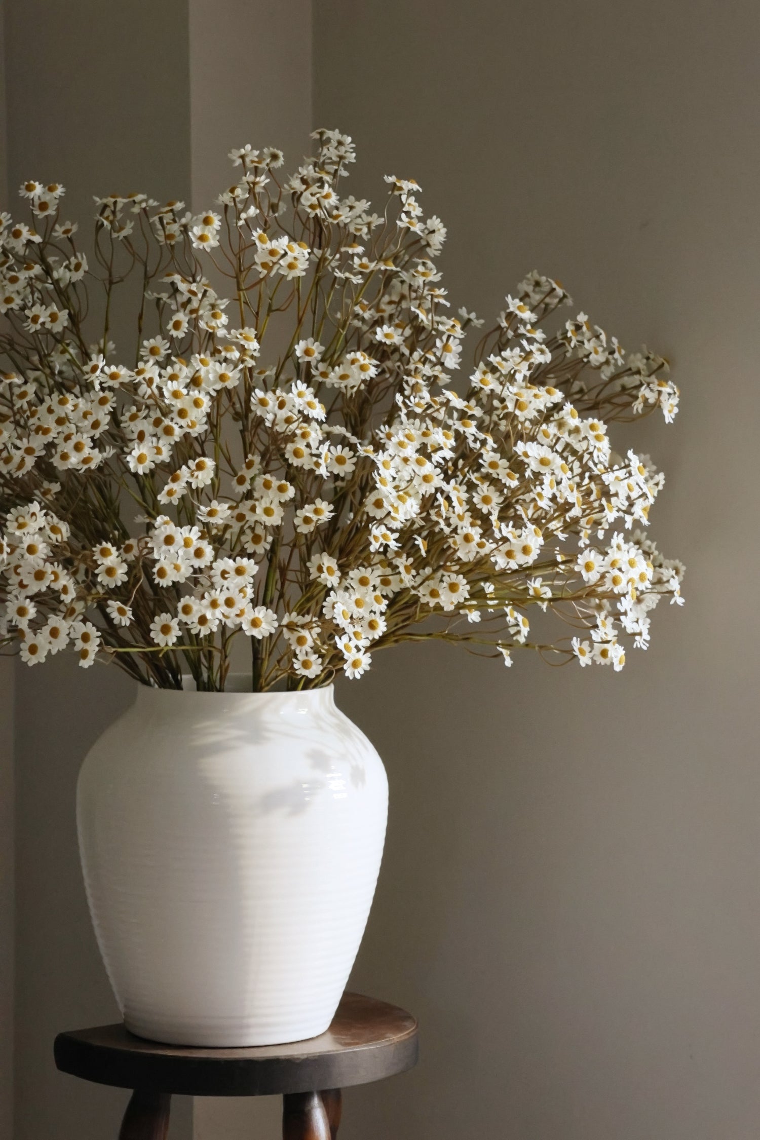 White vase with white daisyflowers on a wooden stool against a plain wall