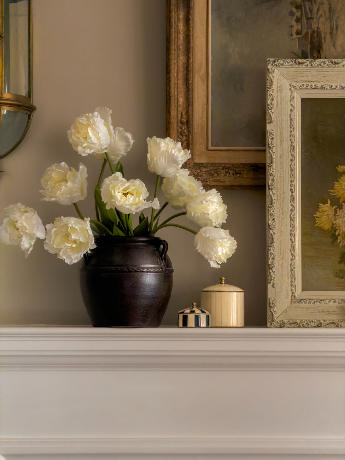 Vase of artificial white tulips flowers on a mantelpiece with decorative items and framed artwork.