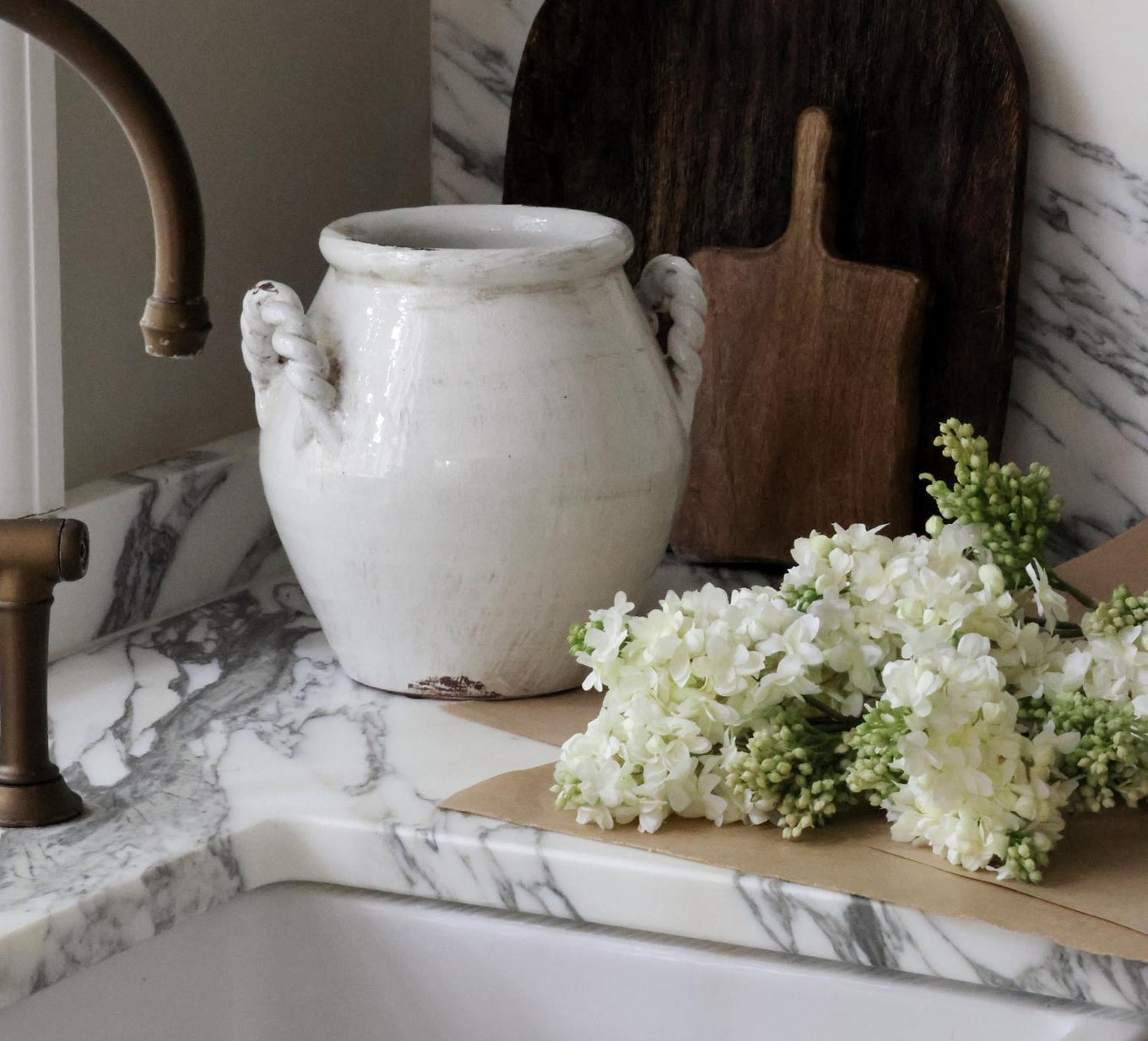 White distressed vase with faux white lilac stems laying alongisde on a marble worktop