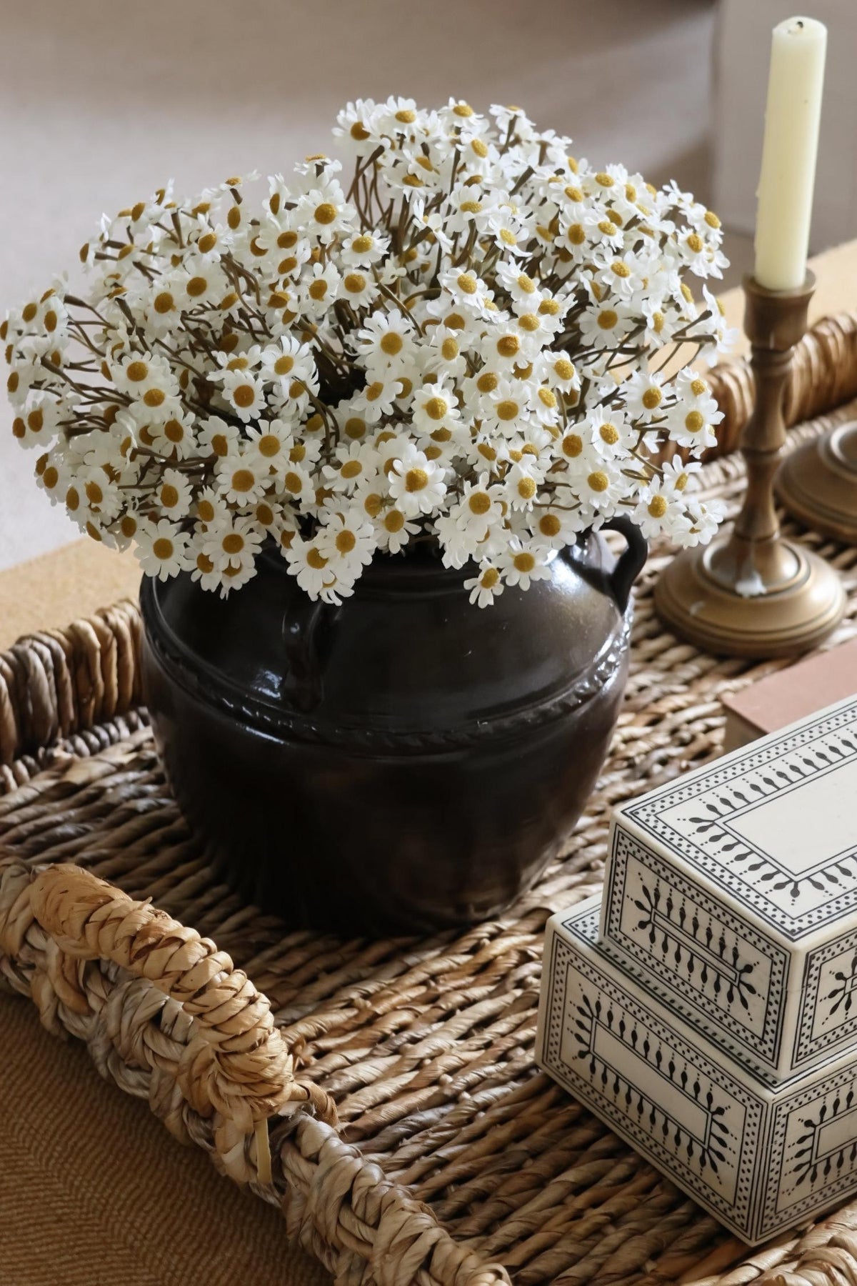 white dasies in a brown pot on a woven tray with a candle stick and decorative box.