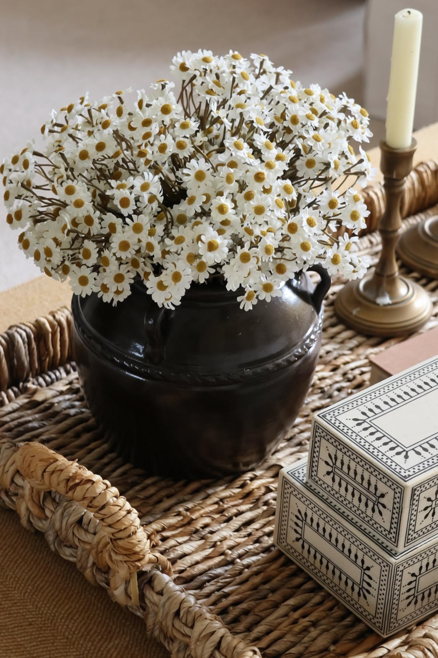 white dasies in a brown pot on a woven tray with a candle stick and decorative box.