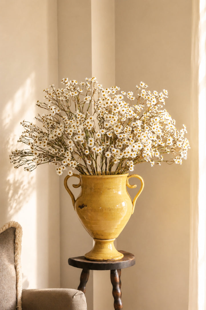 Yellow vase with white daisy flowers on a small wooden table against a beige wall.