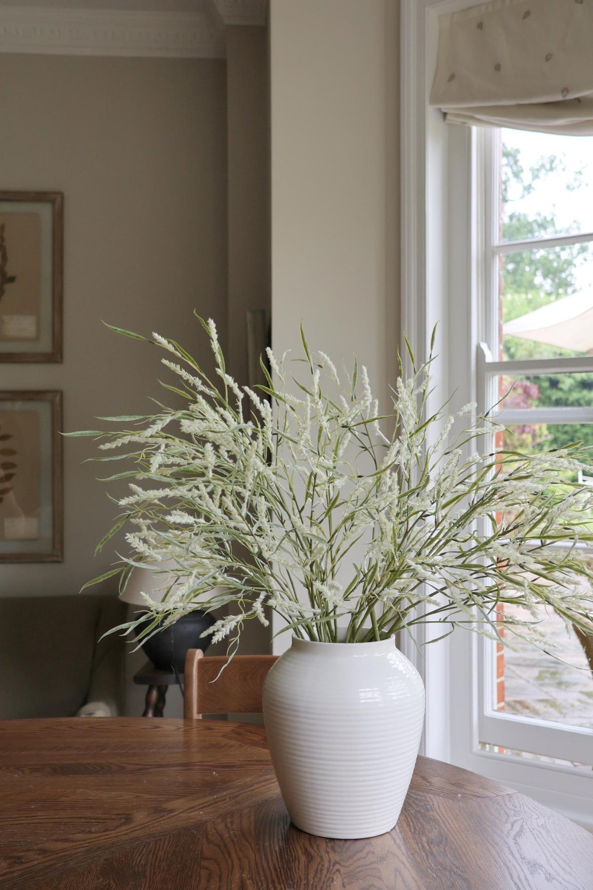 faux wild grass stems in white cermaic vase on wooden table 
