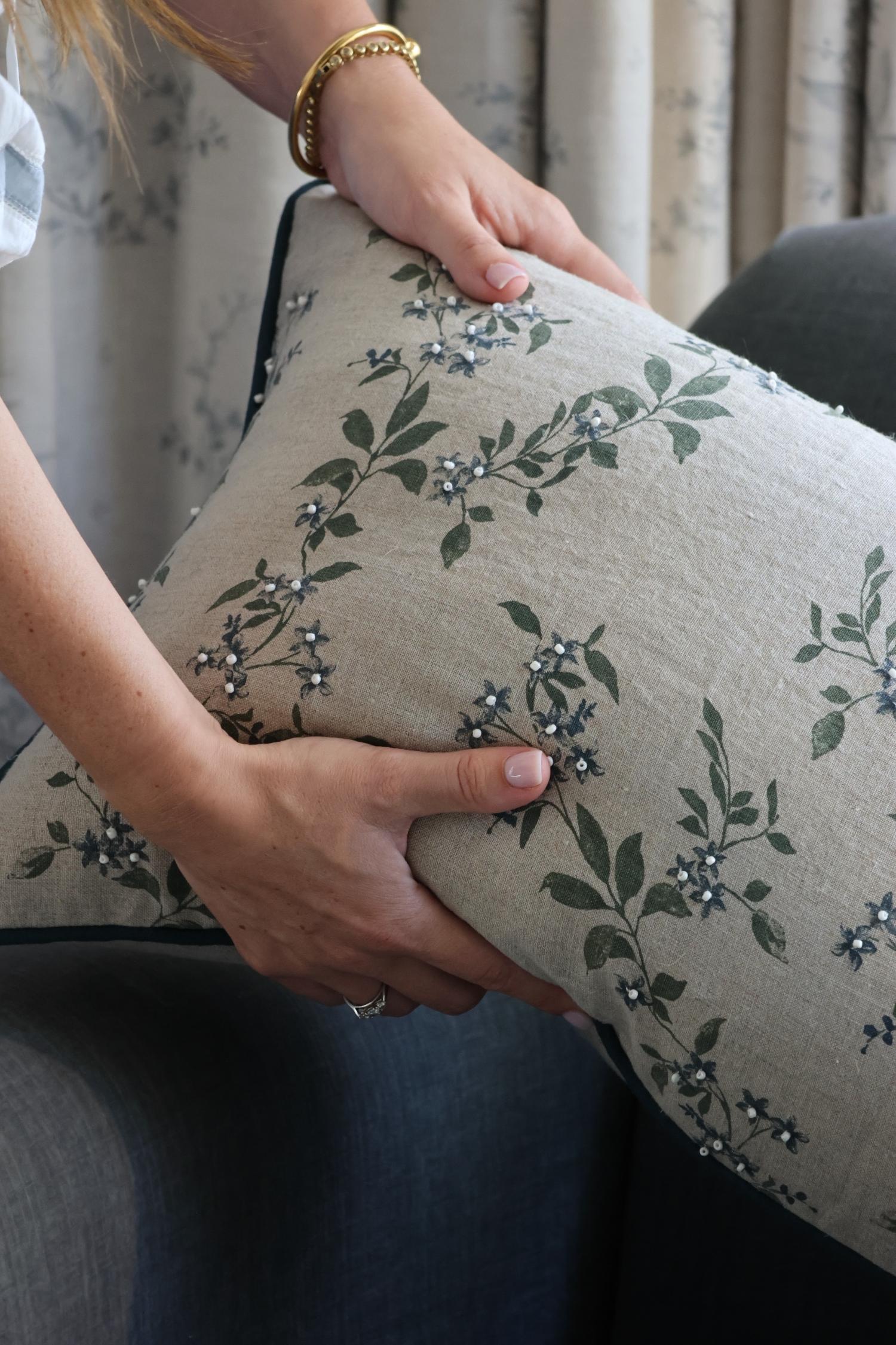 Person holding a floral-patterned cushion with green leaves and blue flowers with blue piping.